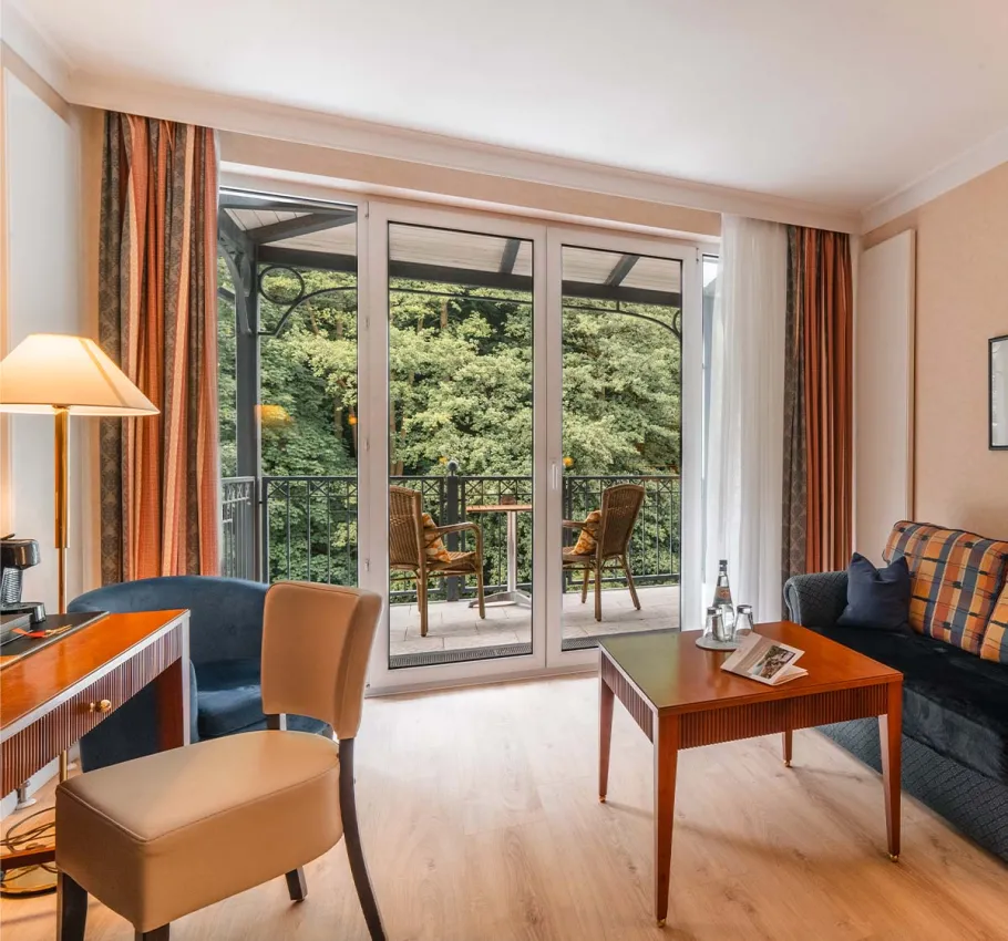 Granitz Deluxe Zimmer im Grand Hotel Binz mit großer Fensterfront, Sofa, Tisch und Balkon mit Blick ins Grüne.