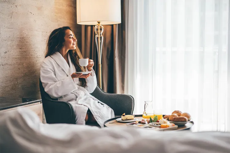 Woman in bathrobe enjoying coffee with room service breakfast in hotel room